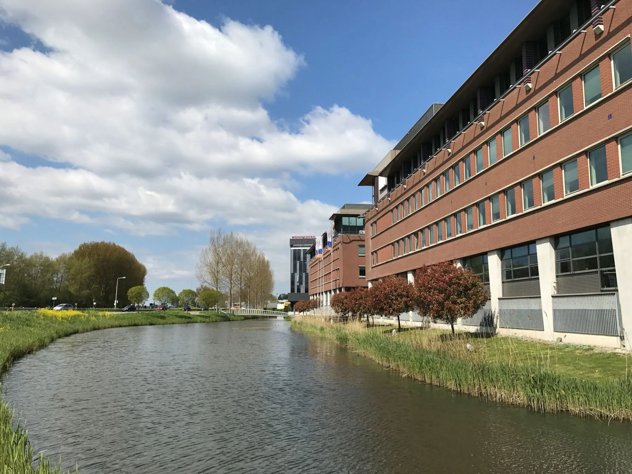 Kantoorpand aan de Tielweg 30 in Gouda met een waterpartij en bomen op de voorgrond onder een blauwe lucht met wolken.