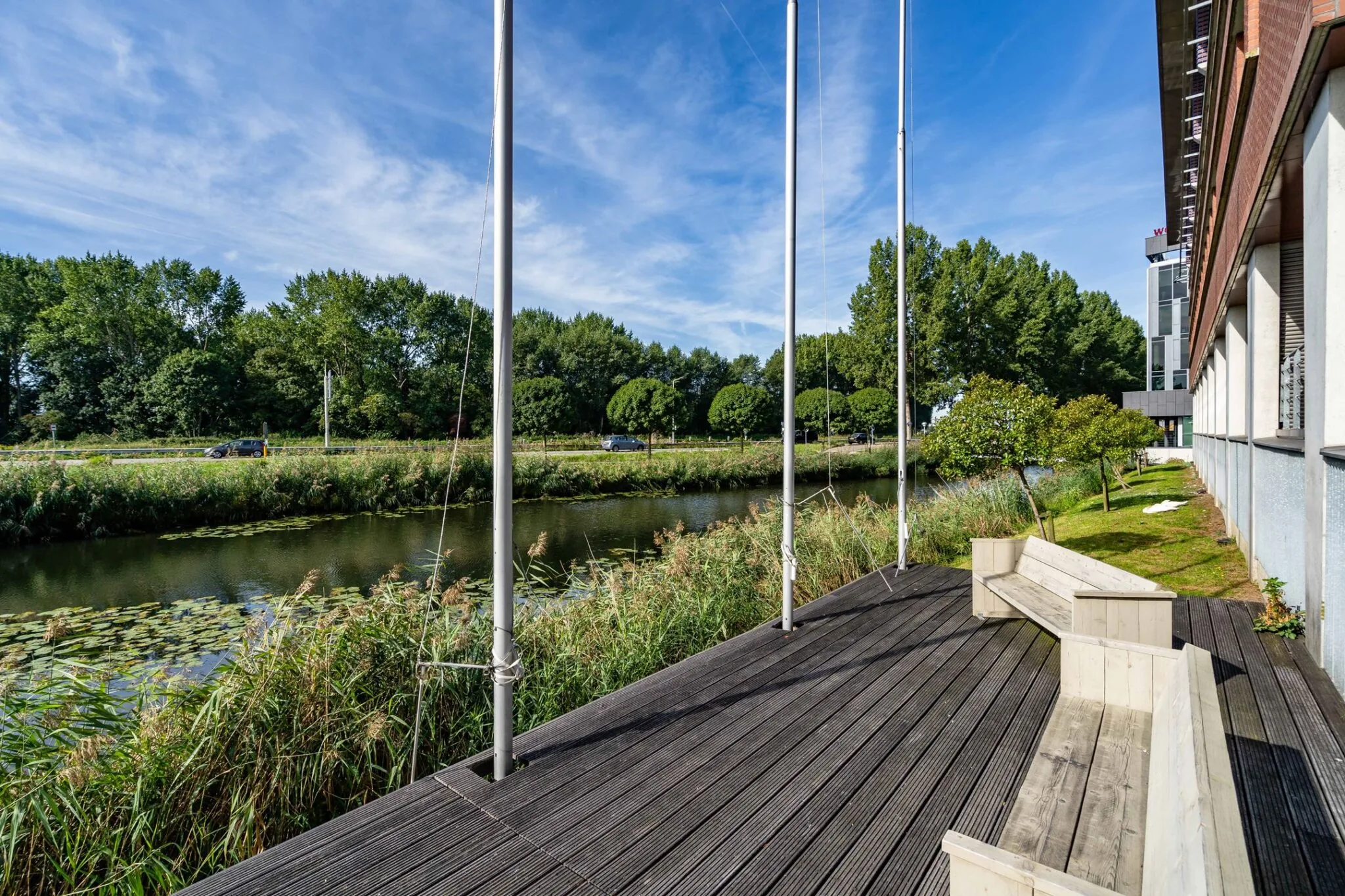 Zicht op het houten terras met zitbanken aan het water bij Tielweg 30, naast een modern kantoorpand en omliggend groen.