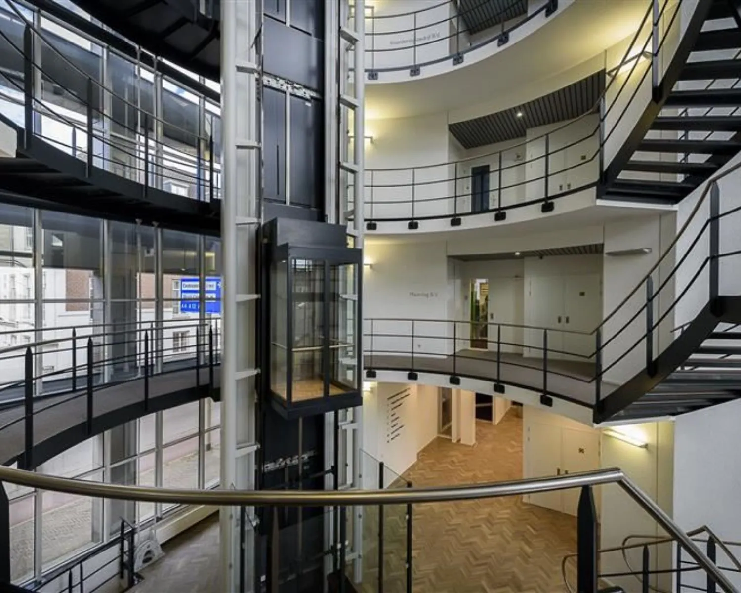 Modern trappenhuis met glazen lift en meerdere verdiepingen met metalen balustrades.