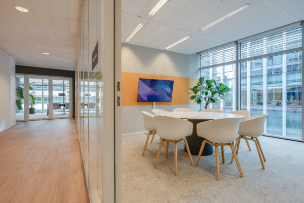 Moderne vergaderruimte met een ronde tafel, stoelen, een groot scherm en een plant, in een lichte kantooromgeving.