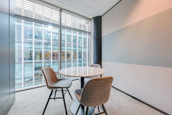 Een moderne vergaderruimte met een ronde tafel en drie stoelen, met uitzicht op een glazen kantoorgebouw.