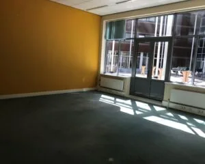 Leeg kantoor met gele muur en grote ramen aan de tuinzijde van Burgemeester van Karnebeeklaan 14 in Den Haag.