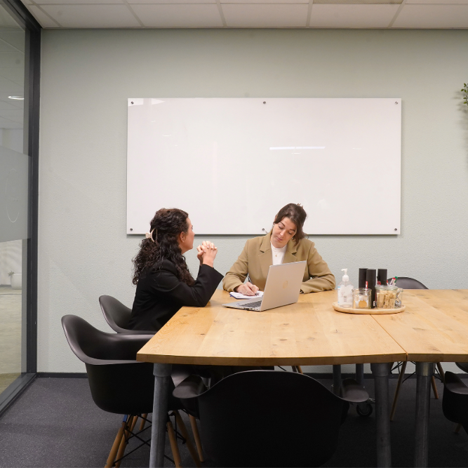 Twee vrouwen hebben een overleg in een vergaderruimte met een houten tafel en een laptop.