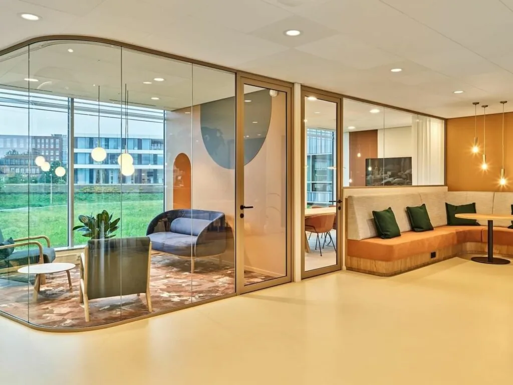 Modern ingerichte kantoorlounge aan de Van Deventerlaan 30-40 met glazen vergaderruimte en zithoek met oranje banken en hanglampen.