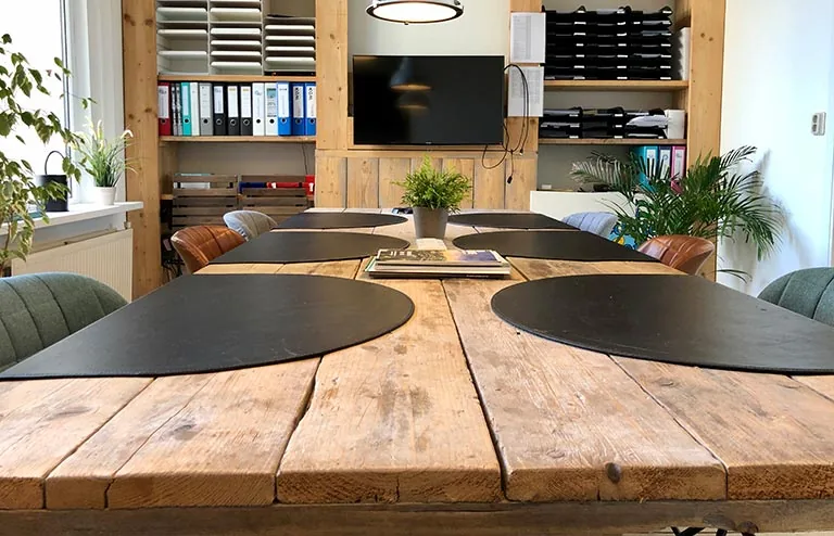 Vergaderruimte aan de Van der Takstraat 8 met een grote houten tafel, zwarte placemats, planten en een tv-scherm aan de muur.