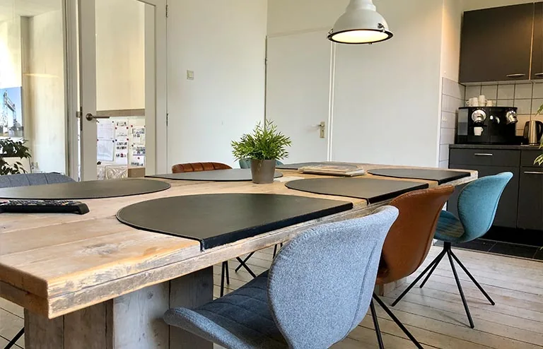 Vergaderruimte met houten tafel, gekleurde stoelen en een kitchenette aan de Van der Takstraat 8.