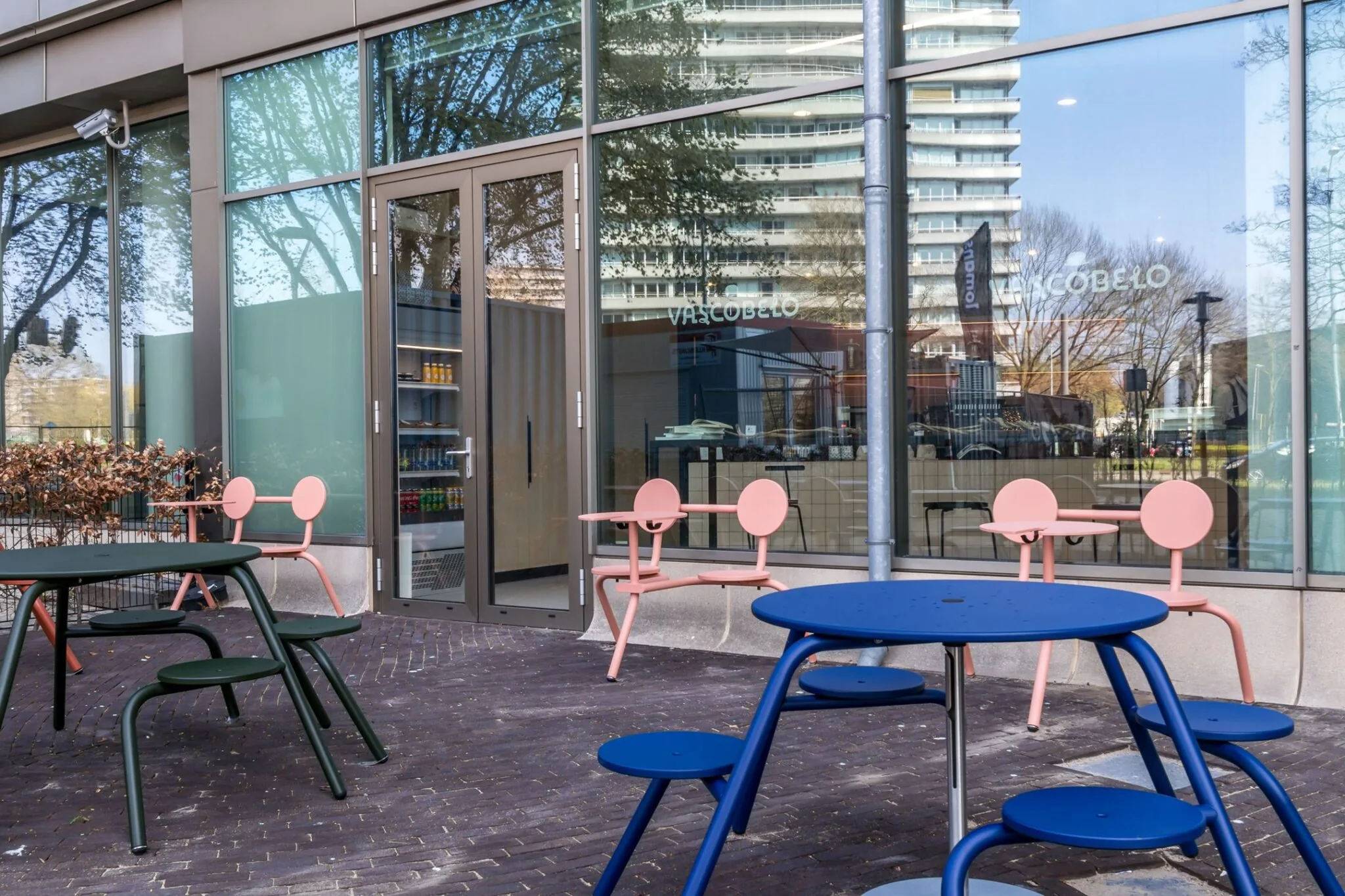 Terras met kleurrijke moderne stoelen en tafels voor de gevel van café Vascobelo aan de Vliegend Hertlaan 21.