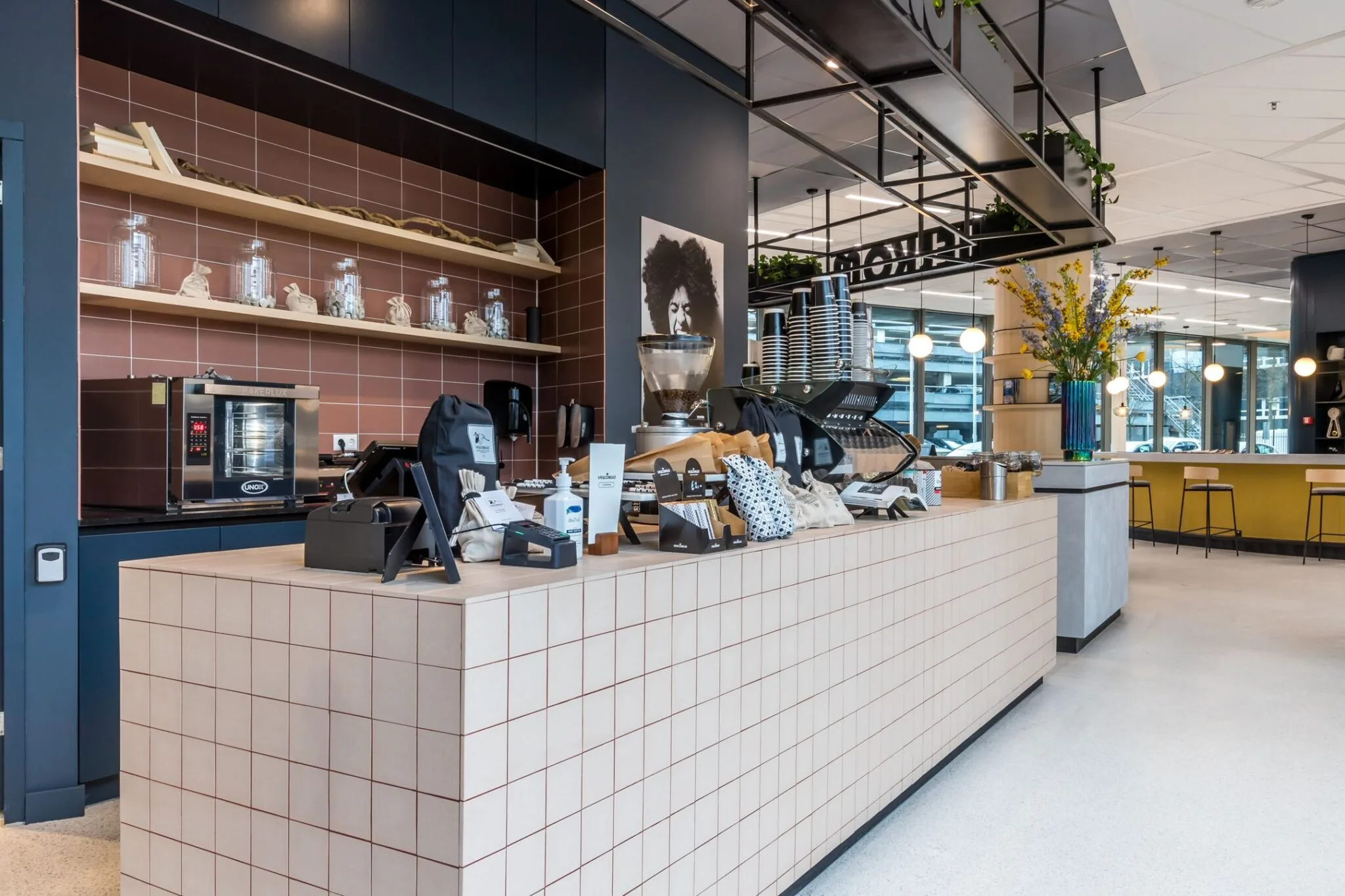 Moderne koffiebar met een betegelde counter, koffiemachine, diverse producten en zitgedeelte in een lichte, open ruimte aan de Vliegend Hertlaan 21.