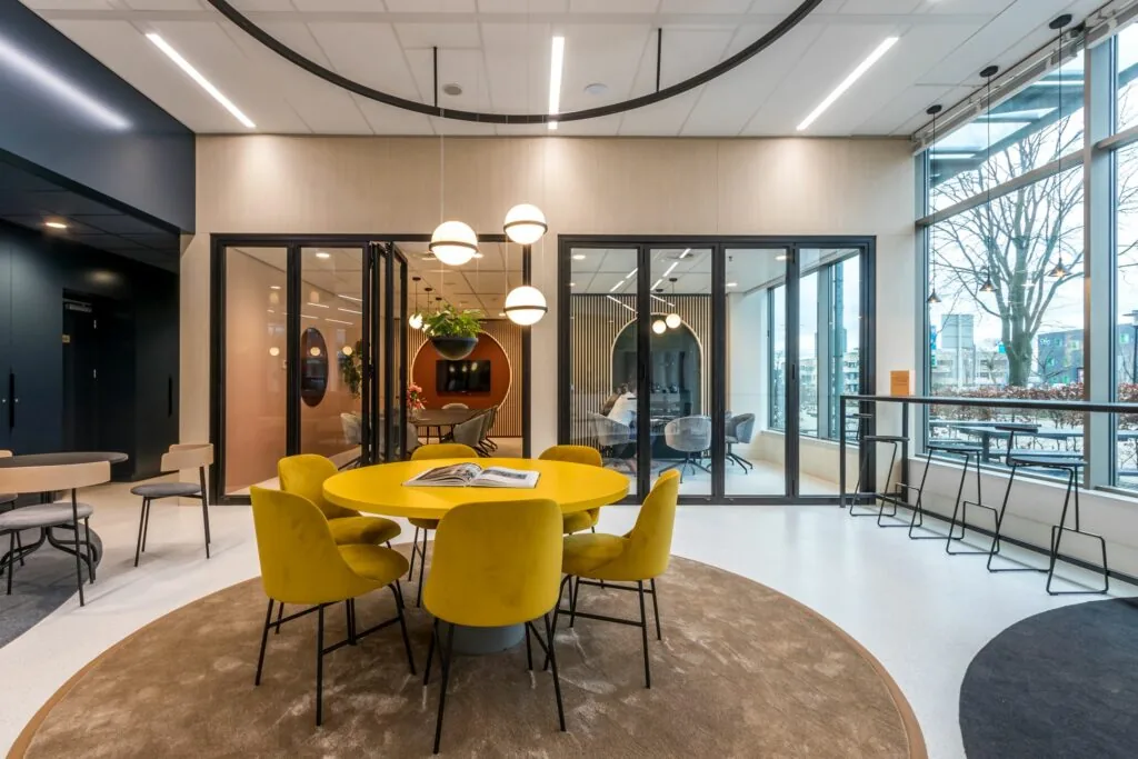 Moderne kantoorruimte aan de Vliegend Hertlaan 21 met gele stoelen rond een ronde tafel en glazen vergaderruimtes op de achtergrond.