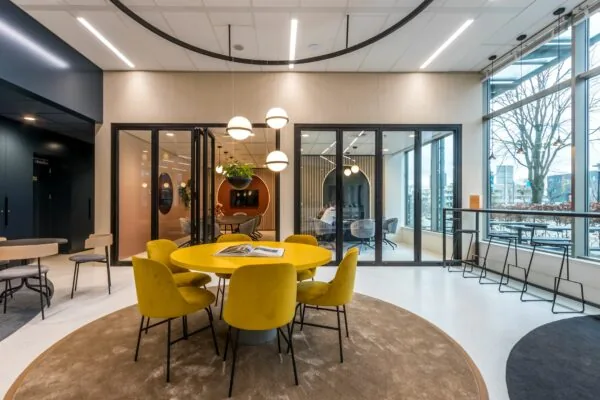 Moderne kantoorruimte aan de Vliegend Hertlaan 21 met gele stoelen rond een ronde tafel en glazen vergaderruimtes op de achtergrond.