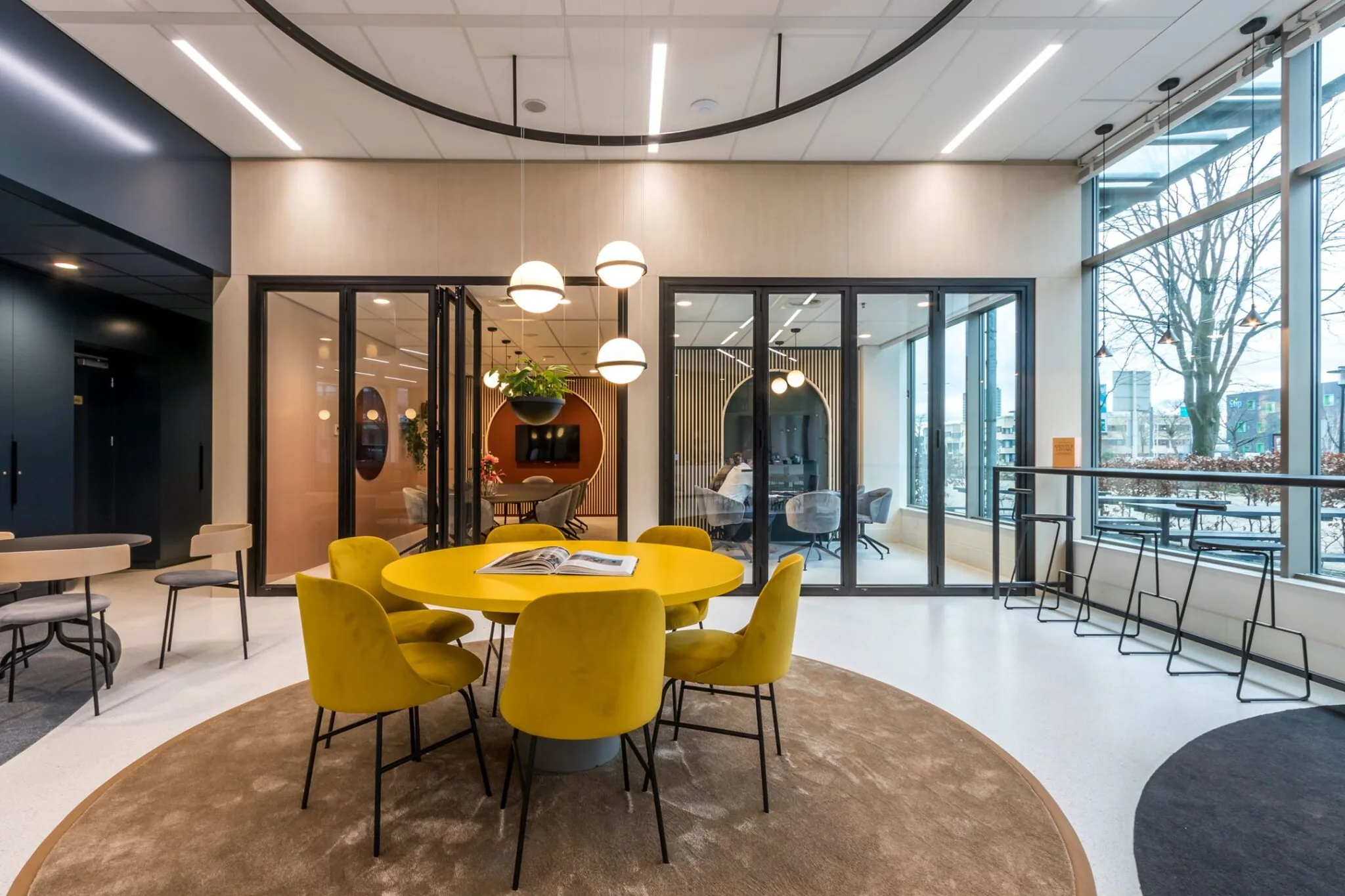 Moderne kantoorruimte aan de Vliegend Hertlaan 21 met gele stoelen rond een ronde tafel en glazen vergaderruimtes op de achtergrond.