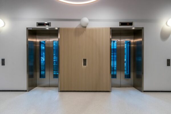 Twee gesloten liften met roestvrijstalen deuren in de hal van Vliegend Hertlaan 21, met een licht houten tussenwand.