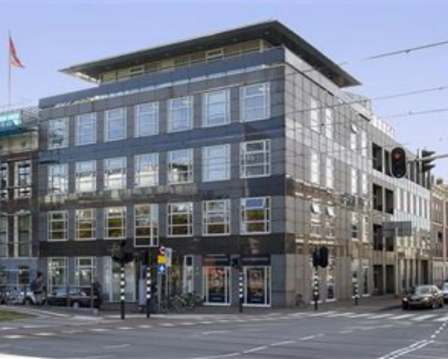 Voorkant van een modern kantoorgebouw met veel ramen op een hoek van een kruispunt in de stad.