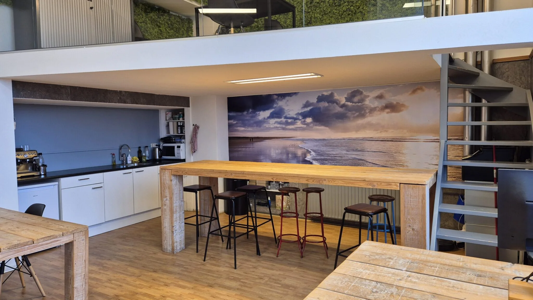 Kantoorkeuken in WG-plein 100 unit 478 met houten tafels, barkrukken, strandfotobehang en trap naar vide.