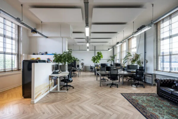 Modern kantoorinterieur met houten visgraatvloer, bureaus, planten en grote ramen met jaloezieën.