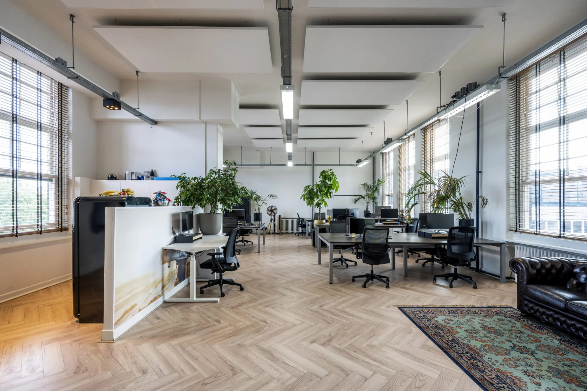 Modern kantoorinterieur met houten visgraatvloer, bureaus, planten en grote ramen met jaloezieën.