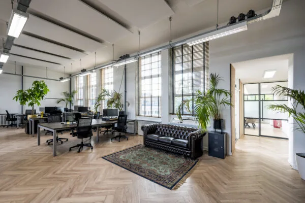 Modern kantoor met houten visgraatvloer, grote ramen, bureaus met computers en een donkerbruine leren chesterfieldbank.