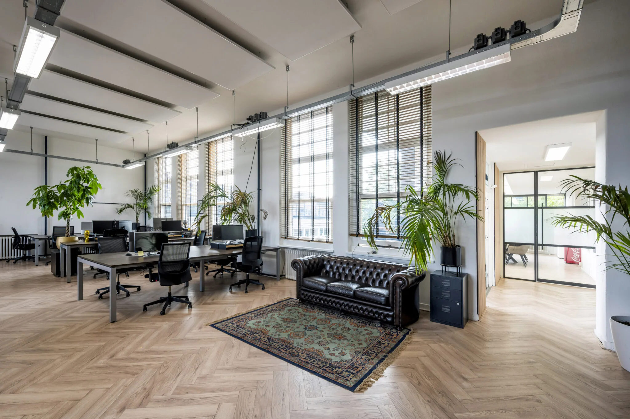 Modern kantoor met houten visgraatvloer, grote ramen, bureaus met computers en een donkerbruine leren chesterfieldbank.