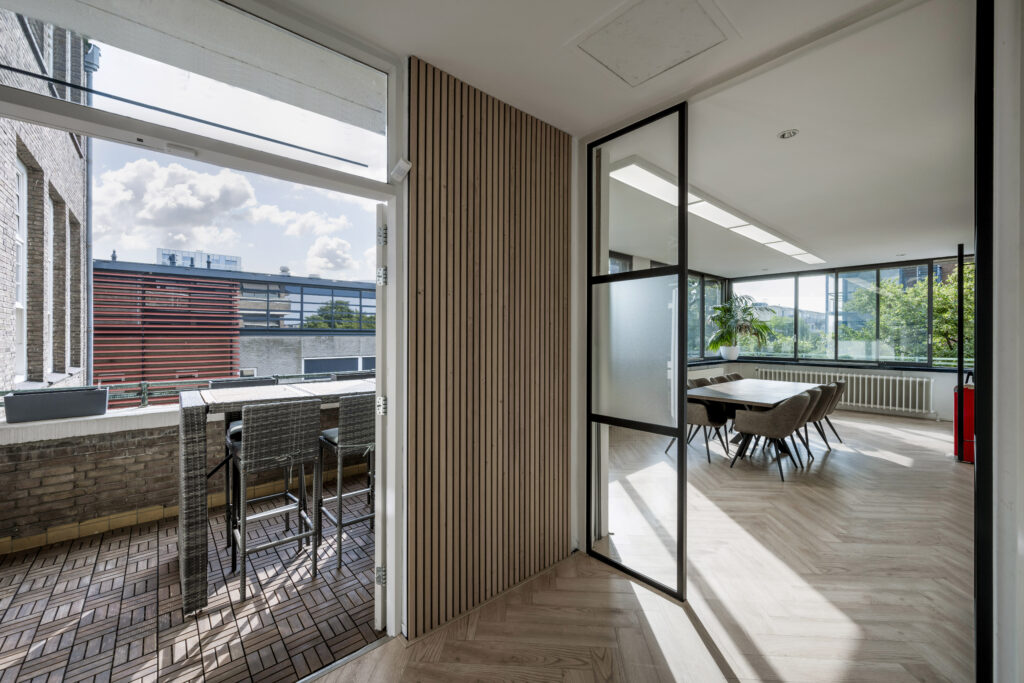Moderne vergaderruimte met grote ramen en een aangrenzend balkon met hoge stoelen en houten vloer.
