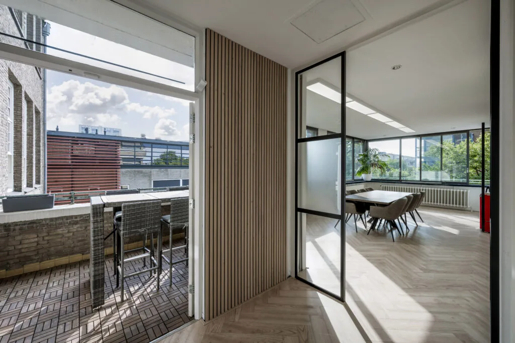 Moderne vergaderruimte met grote ramen en een aangrenzend balkon met hoge stoelen en houten vloer.