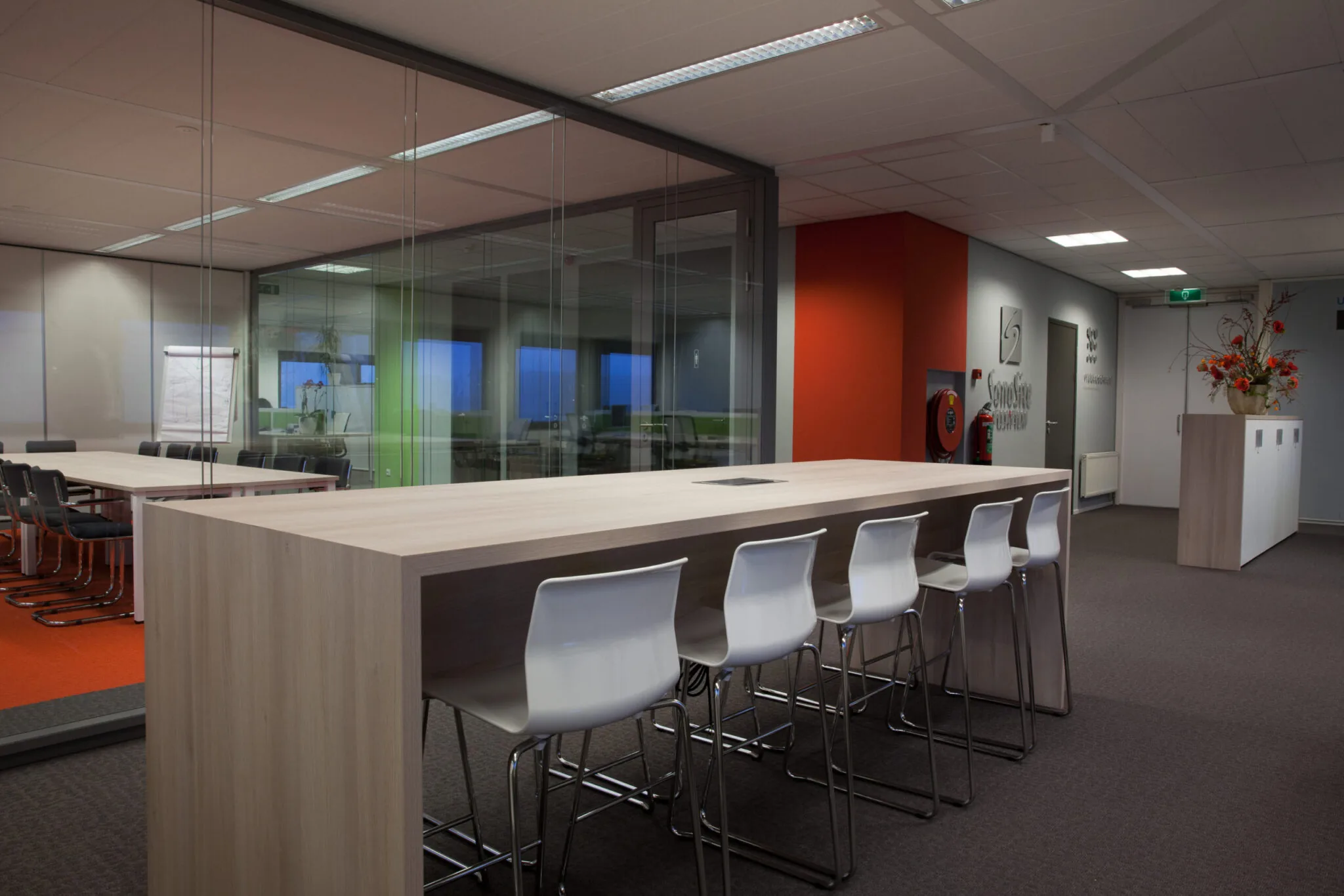 Moderne kantoorruimte met een hoge vergadertafel, witte barkrukken en een glazen vergaderruimte op de achtergrond.