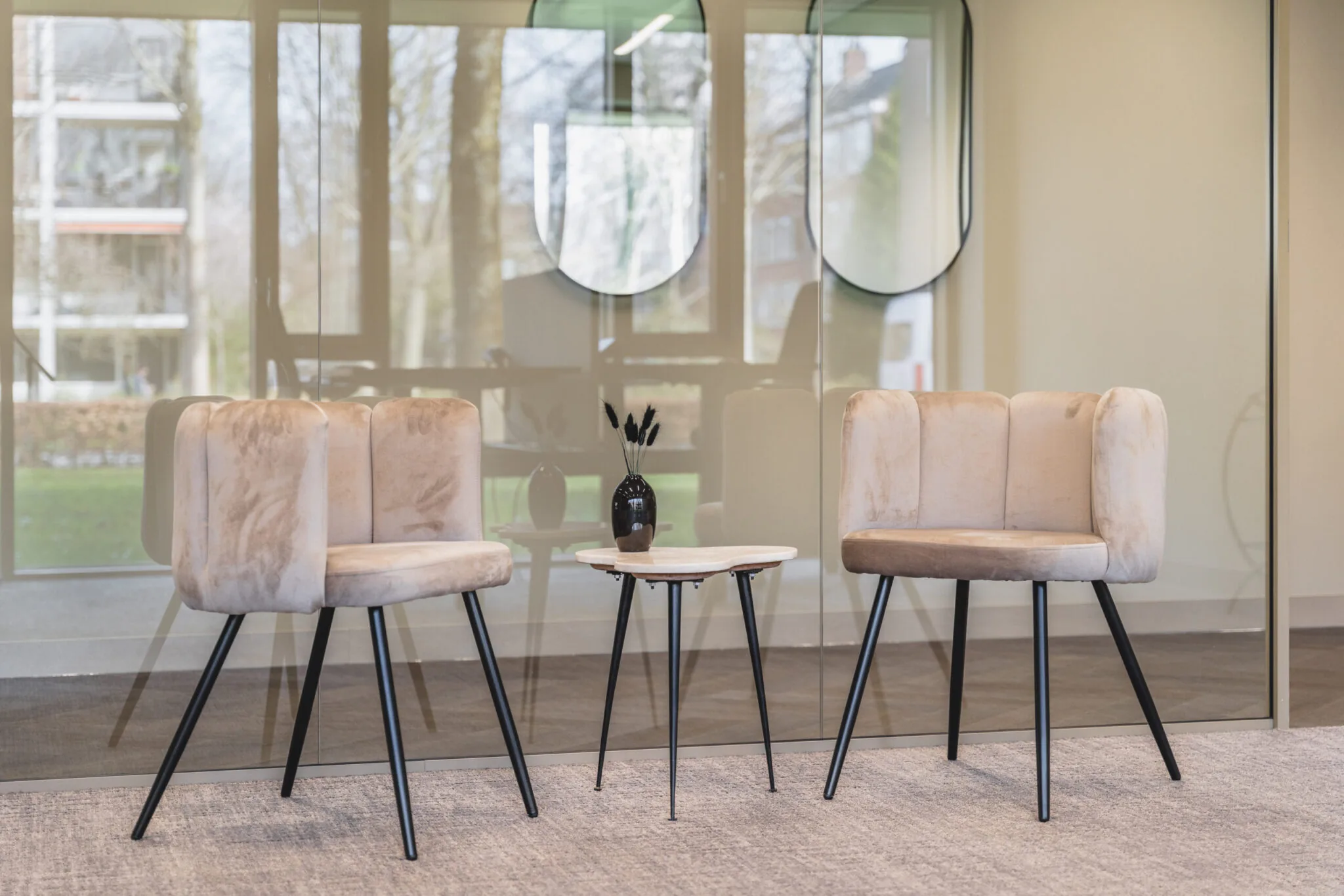 Twee beige fluwelen stoelen met een rond bijzettafeltje ertussen in een moderne wachtruimte bij Walborg.