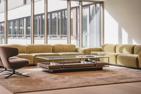 Moderne loungehoek in Walborg met een grote gele hoekbank, glazen salontafel en een bruine draaistoel bij hoge ramen.