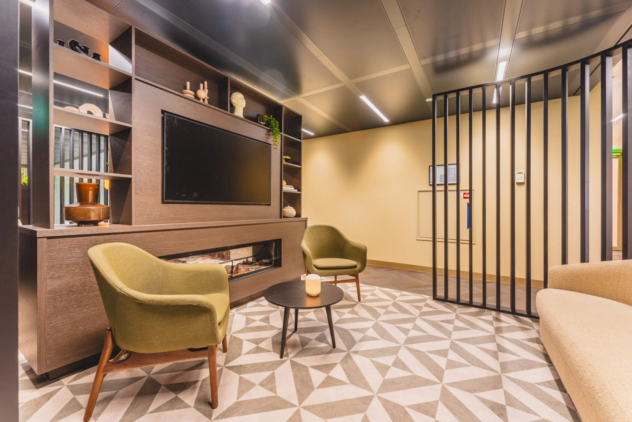 Moderne wachtruimte met twee groene stoelen, een ronde salontafel, een ingebouwde tv en decoratieve houten wandkasten.