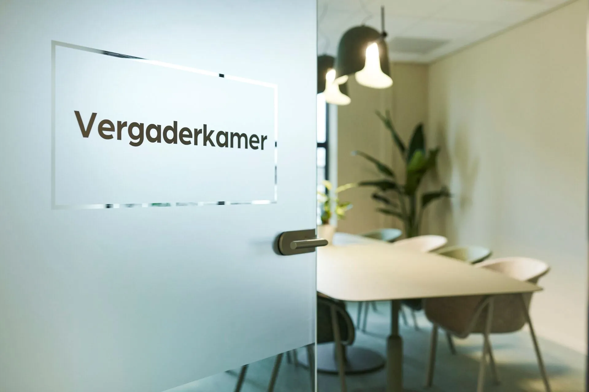 Een moderne vergaderkamer met glazen deur waarop 'Vergaderkamer' staat, ingericht met een ovale tafel, stoelen en hanglampen.