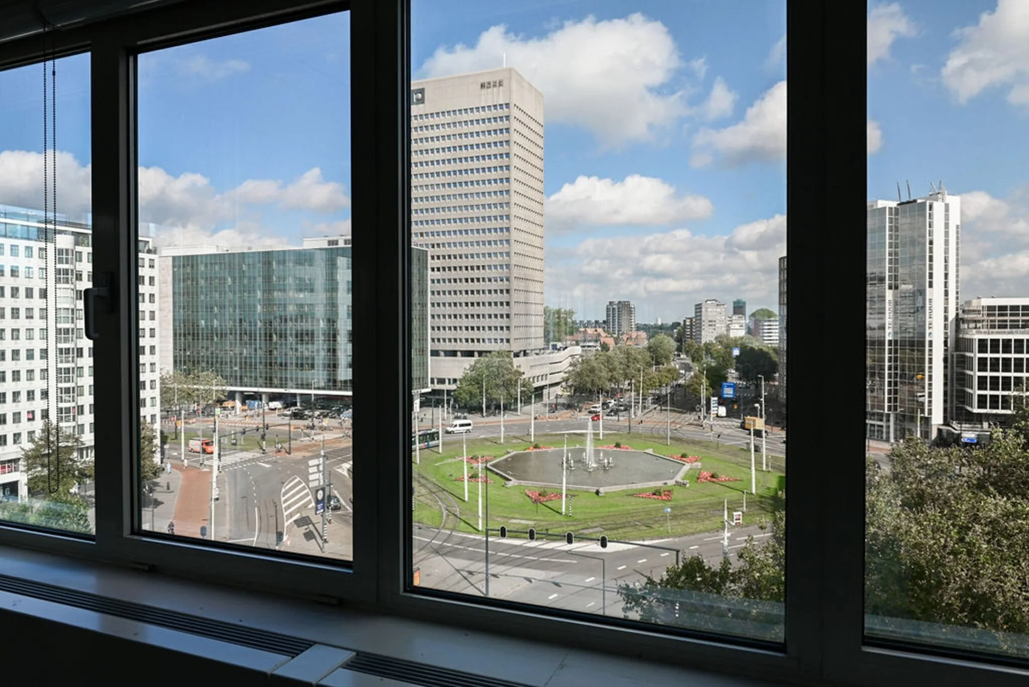 Uitzicht vanuit een raam op het Weena in Rotterdam met een rotonde, fontein en omliggende kantoorgebouwen.