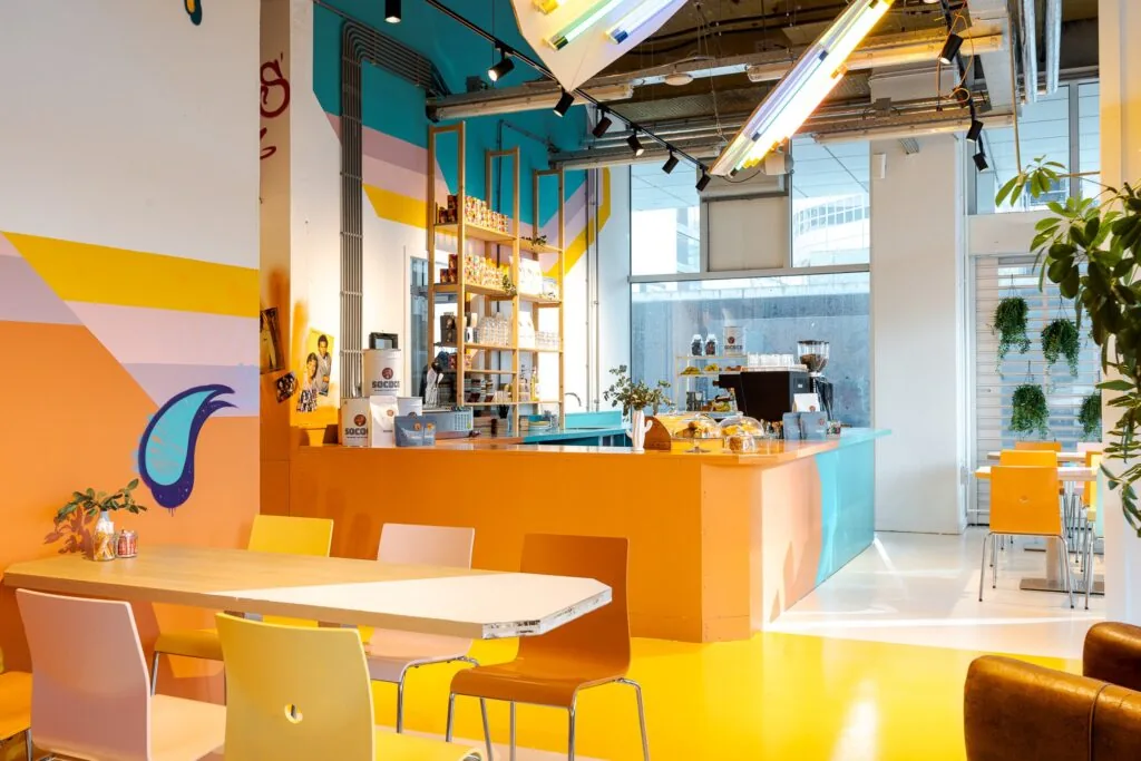 Kleurrijk en modern interieur van een café in Weena 70 met een oranje en turquoise balie, zitplaatsen en grote ramen.