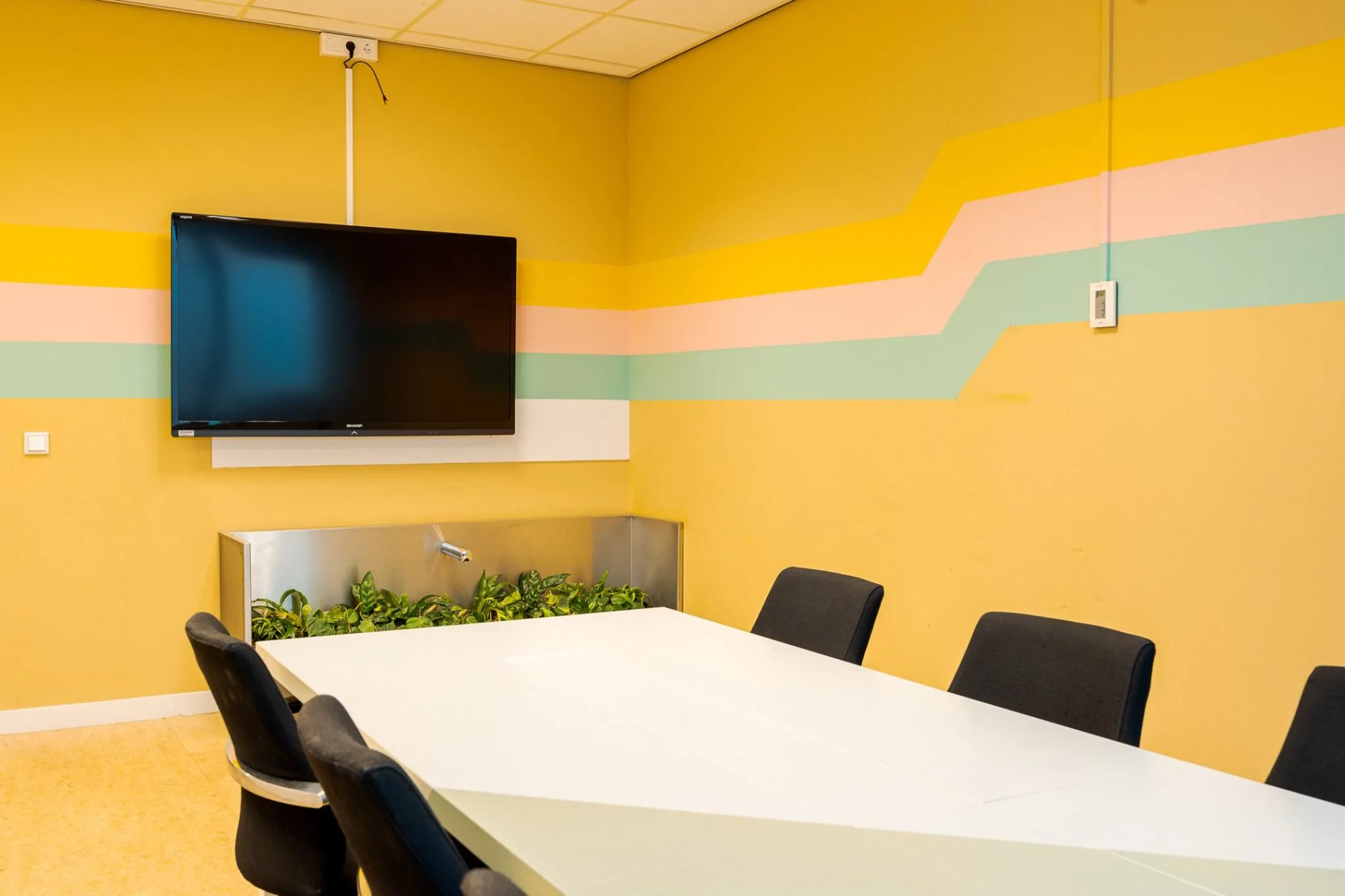 Vergaderruimte in Weena 70 met gele muur, gekleurde wandstrepen, flatscreen en witte tafel met zwarte stoelen.