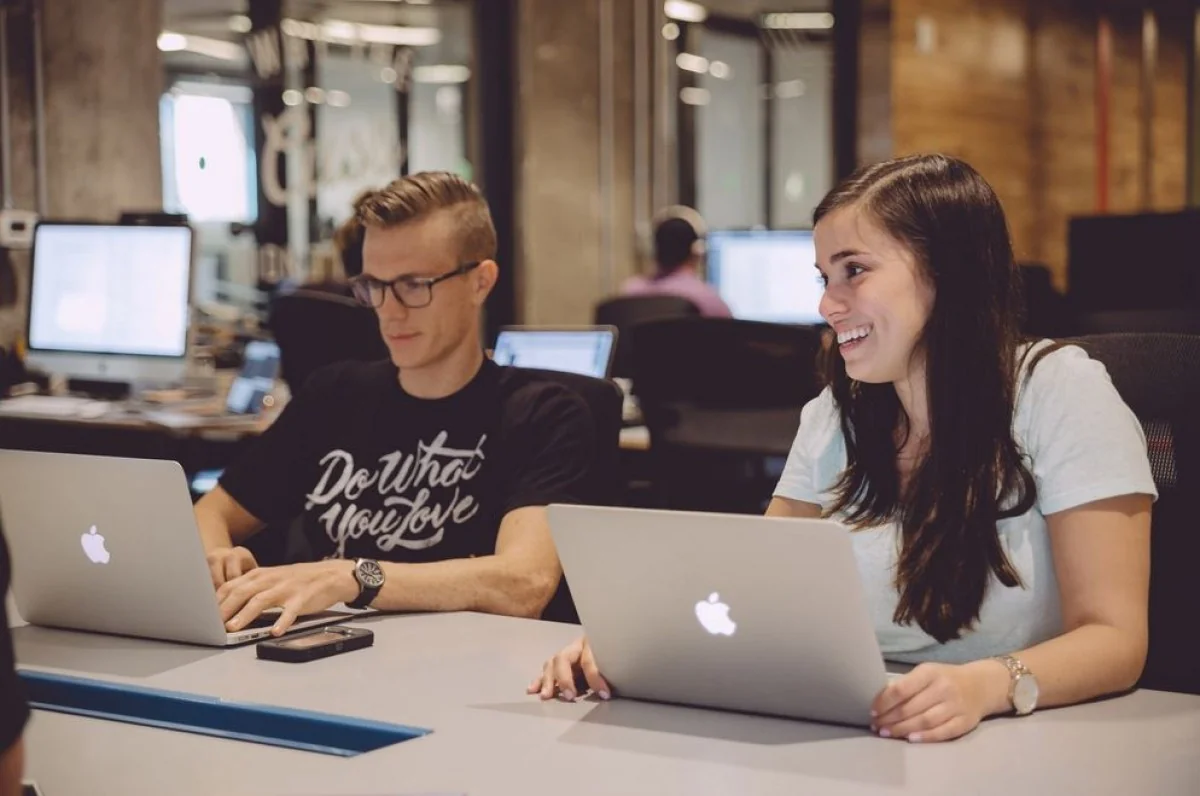 Twee jonge professionals werken lachend op hun laptops in een modern kantoor aan de Weesperstraat.
