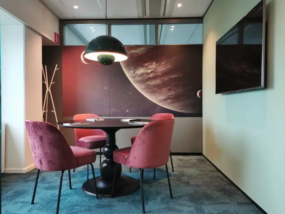 Een vergaderruimte met een ronde tafel, roze stoelen en een muur met een afbeelding van een planeet.