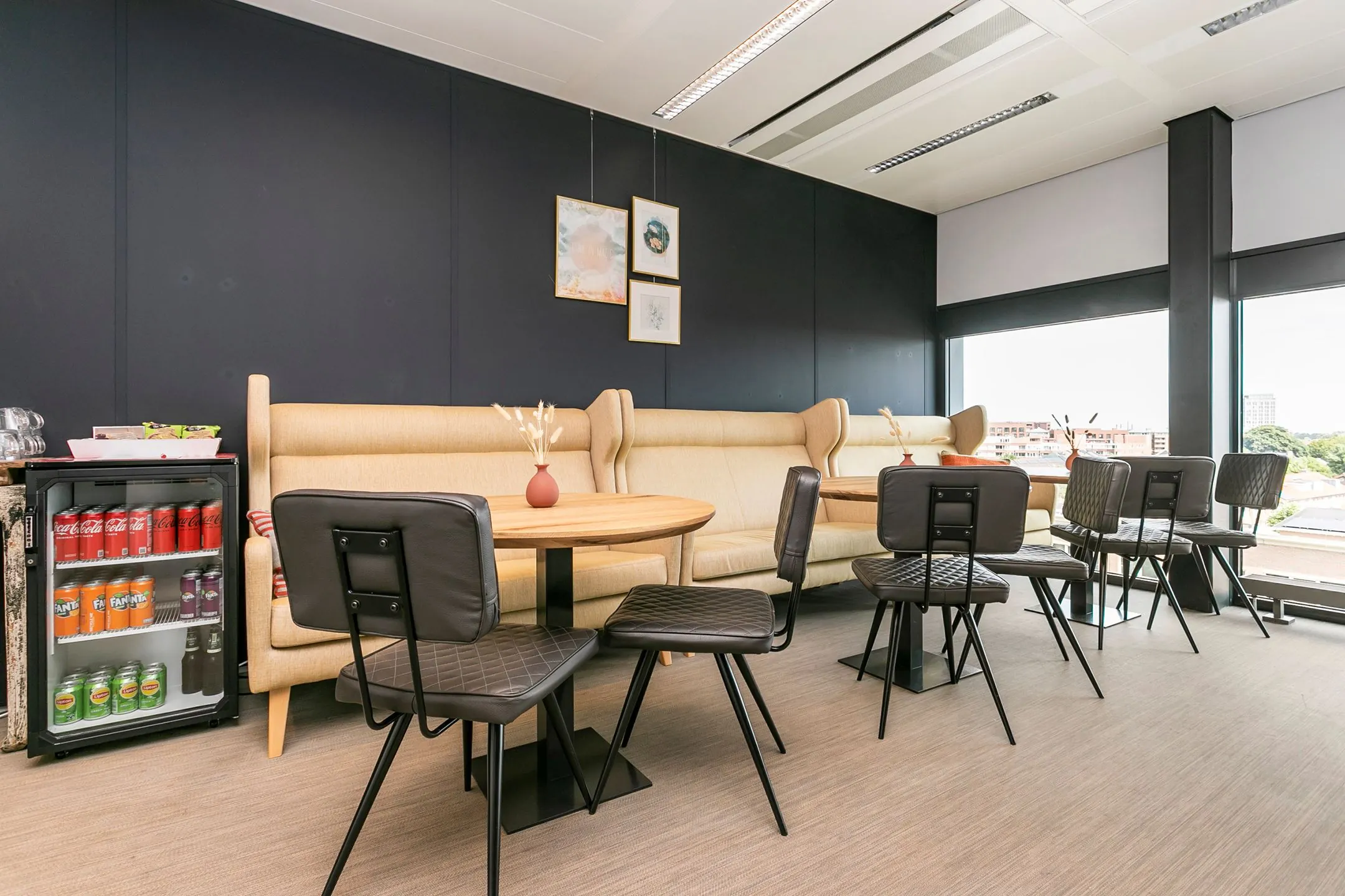 Moderne loungehoek met beige banken, zwarte stoelen, ronde houten tafels en een koelkast met frisdrank in een kantoorruimte aan het Wim Duisenbergplantsoen.