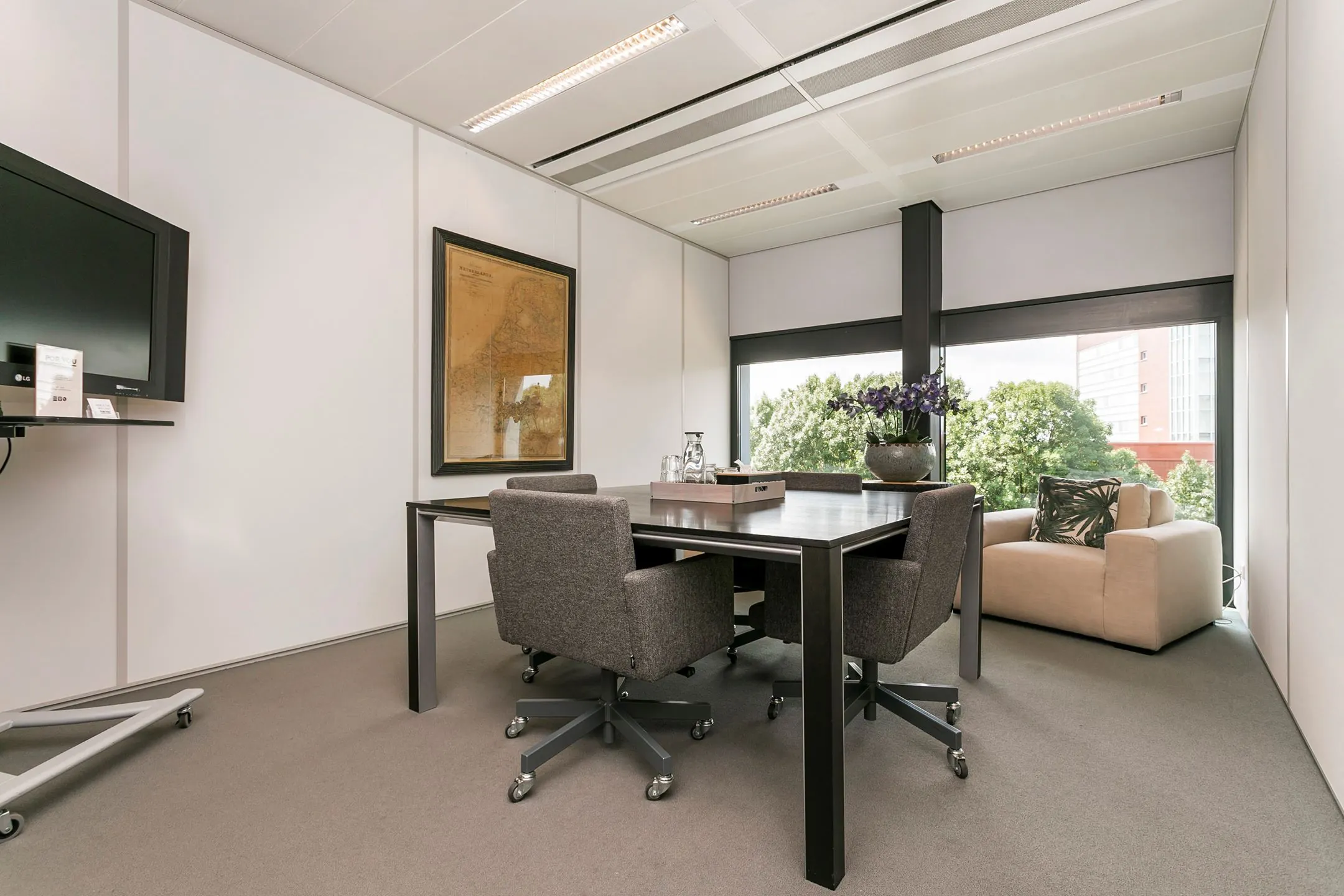 Vergaderruimte met moderne inrichting, een tafel met vier stoelen, een grote plant en uitzicht op bomen en een gebouw aan het Wim Duisenbergplantsoen.