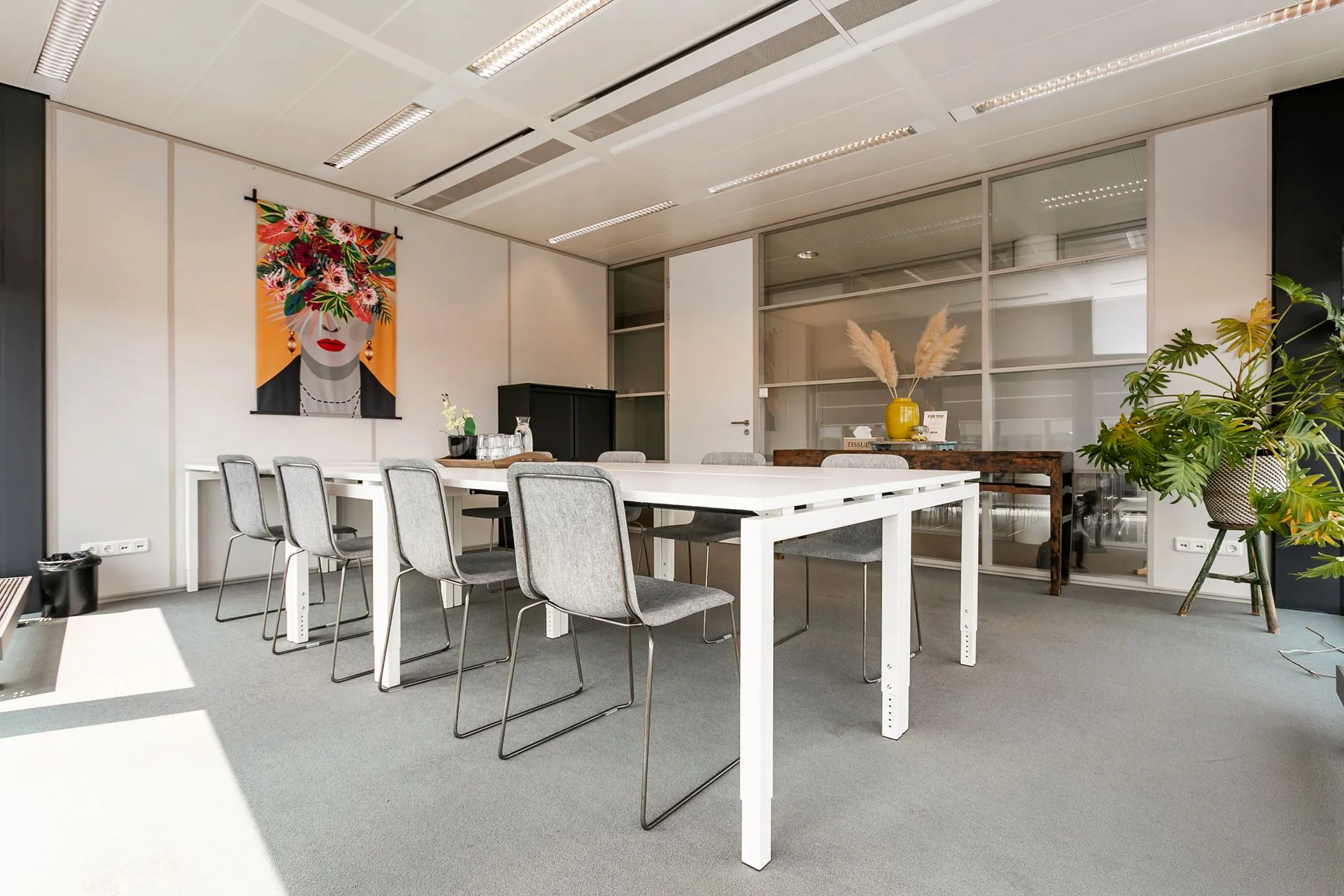 Moderne vergaderruimte aan het Wim Duisenbergplantsoen 31 met een witte tafel, grijze stoelen, kleurrijke wanddecoratie en veel natuurlijk licht.