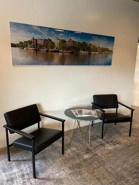 Twee zwarte stoelen met een rond tafeltje ertussen in een wachtruimte, met een wandfoto van Amsterdam op de achtergrond.