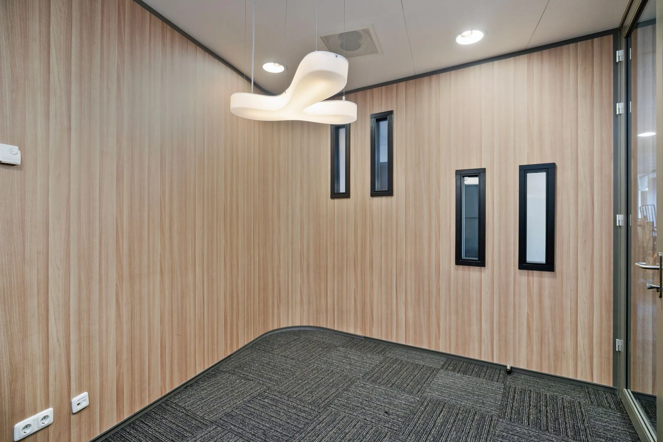 Kleine moderne kantoorruimte met houtlook wanden, drie smalle ramen en een opvallende hanglamp.