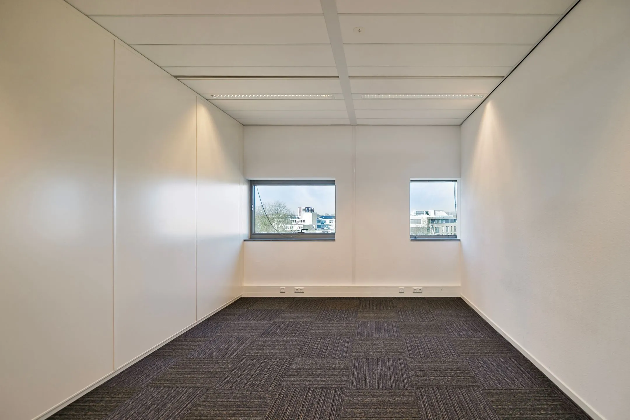 Leeg, smal kantoor met tapijtvloer, witte wanden en twee ramen aan het einde met uitzicht op Zwartewaterallee.