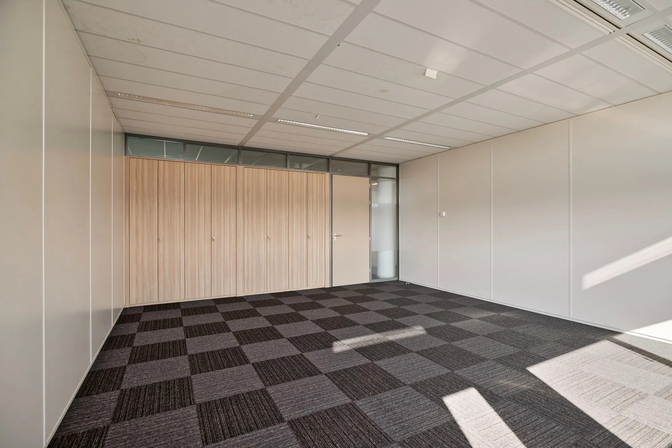 Lege kantoorruimte aan de Zwartewaterallee 56 met tapijttegels, ingebouwde houten kasten en een glazen deur.