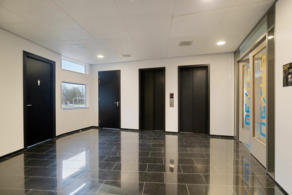 Moderne hal met twee liften, zwarte deuren, glanzende tegelvloer en een glazen toegangsdeur naar Nexus in Zwartewaterallee 56.