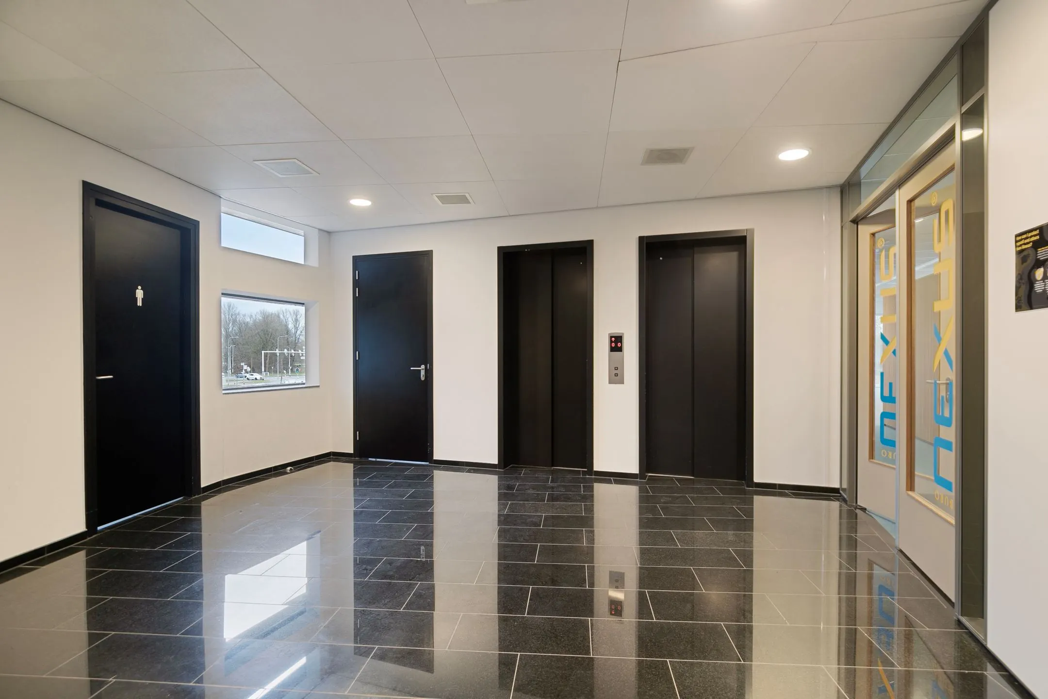 Moderne hal met twee liften, zwarte deuren, glanzende tegelvloer en een glazen toegangsdeur naar Nexus in Zwartewaterallee 56.