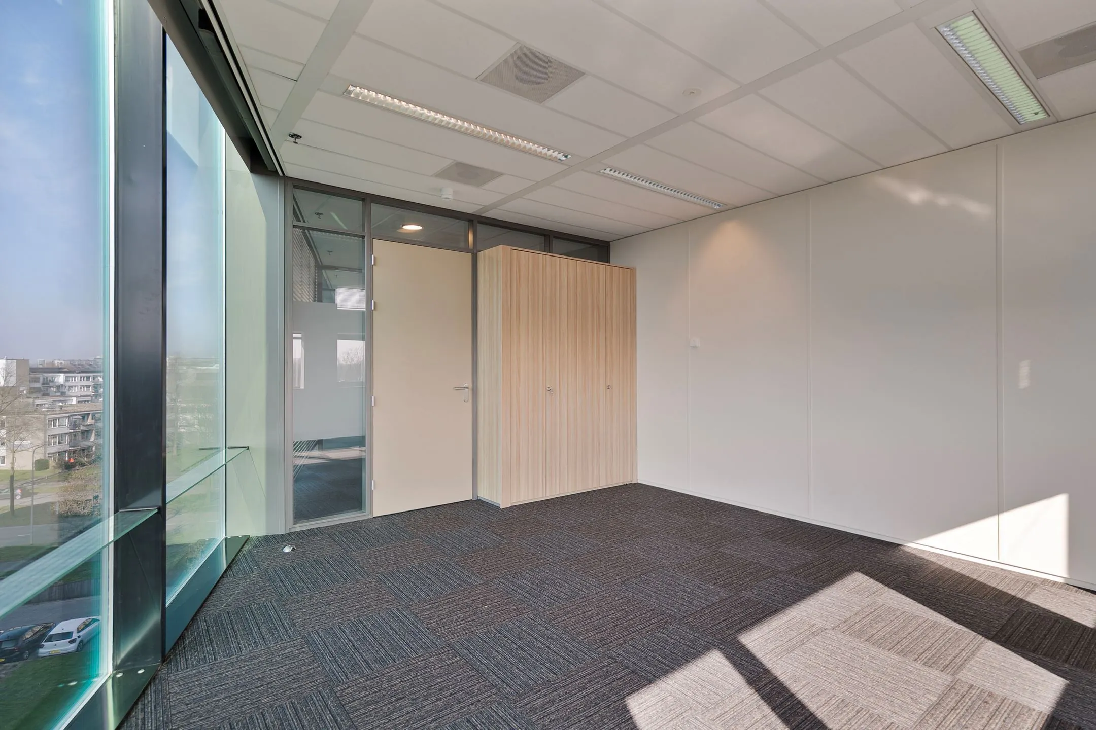 Leeg modern kantoor met glazen buitenwand, grijze tapijttegels en een houten opbergkast aan de Zwartewaterallee 56.