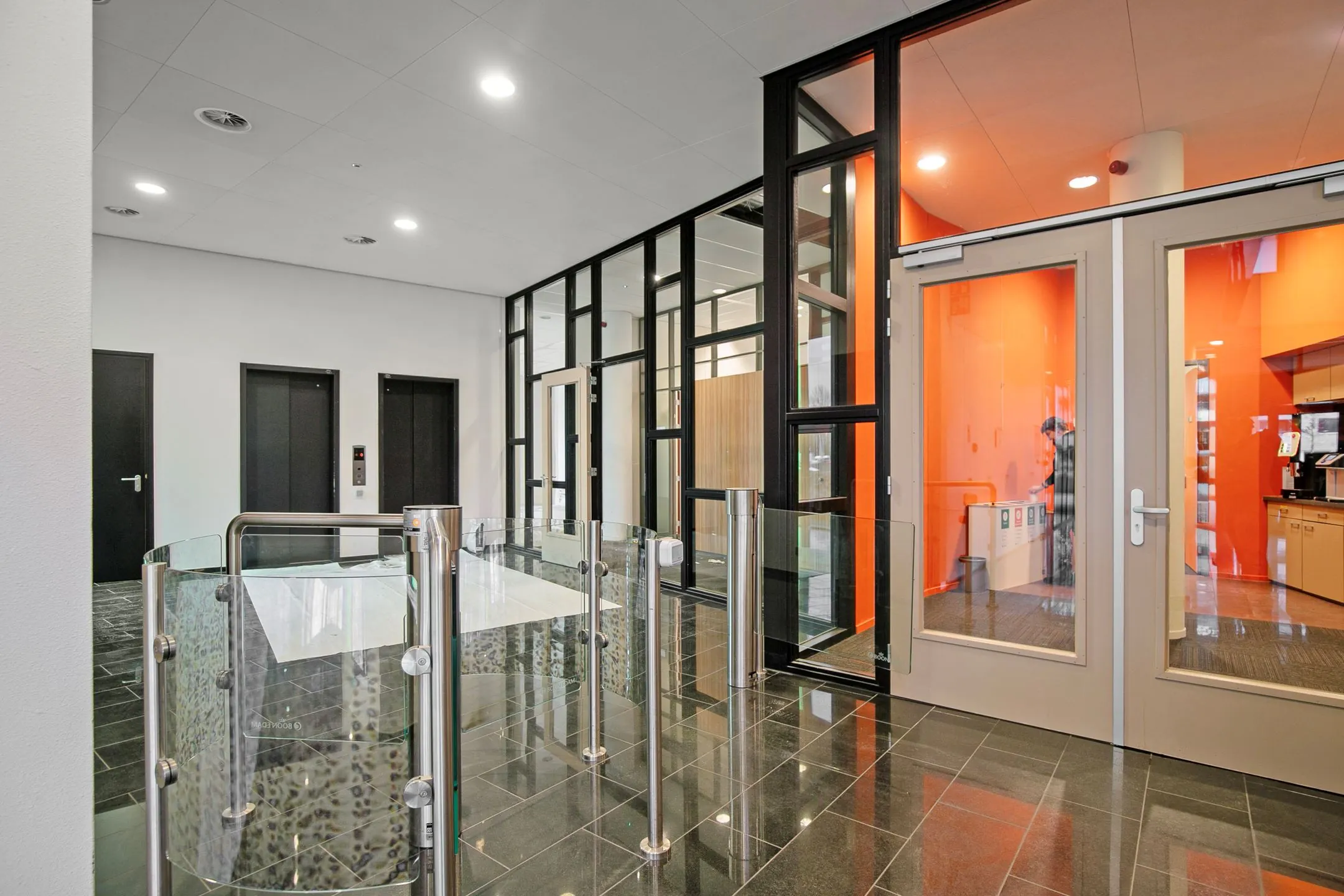 Ingangsgebied van het kantoorgebouw aan de Zwartewaterallee 56 met glazen toegangspoortjes, liften en een feloranje wand in de achtergrond.