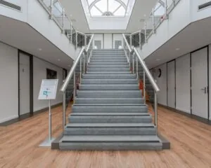Brede trap met grijze treden en metalen leuningen in een modern, licht atrium met houten vloer.