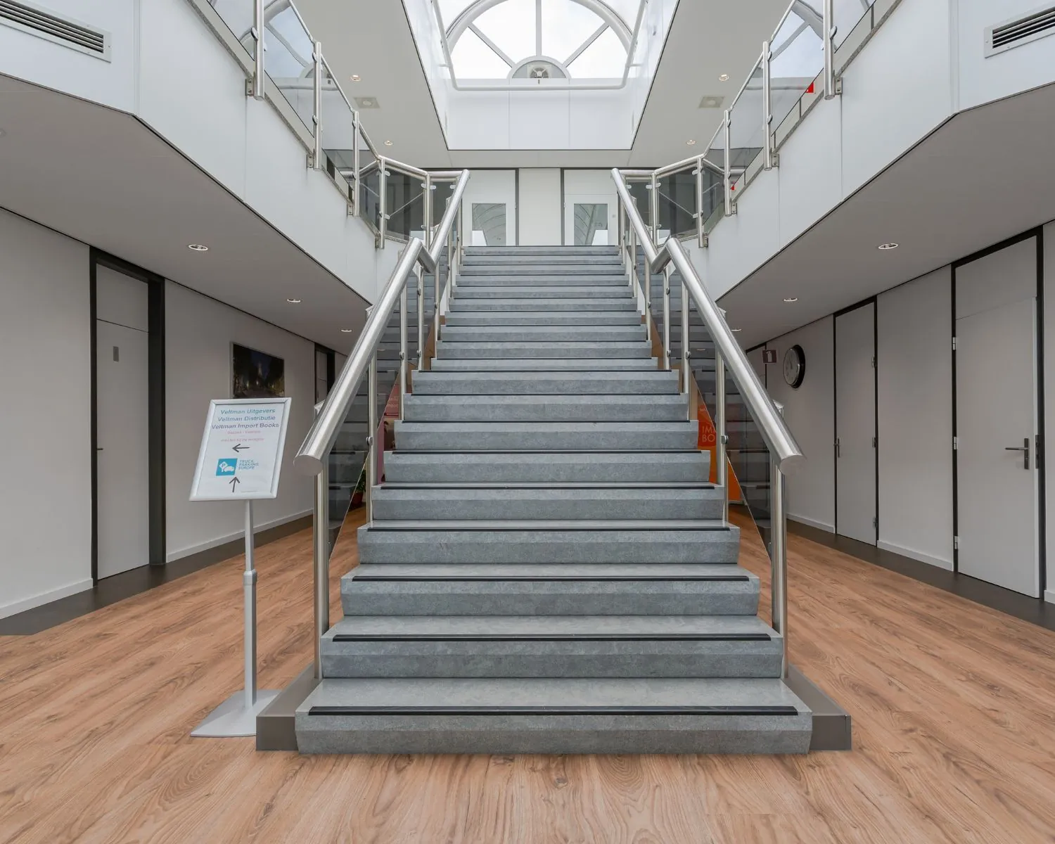 Brede trap met grijze treden en metalen leuningen in een modern, licht atrium met houten vloer.