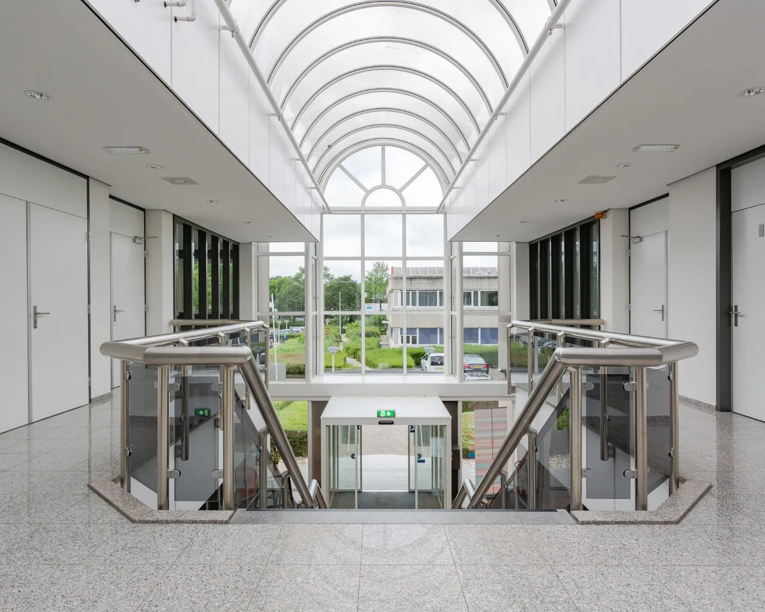 Moderne, lichte kantoorhal met glazen dak, roestvrijstalen trapleuningen en uitzicht op een groen bedrijventerrein.