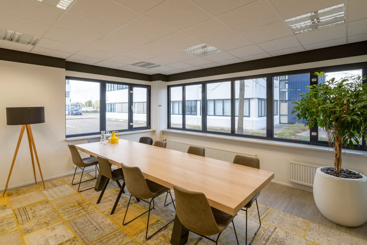 Vergaderruimte met lange houten tafel, acht stoelen, grote ramen en uitzicht op kantoren aan de Atoomweg 400.