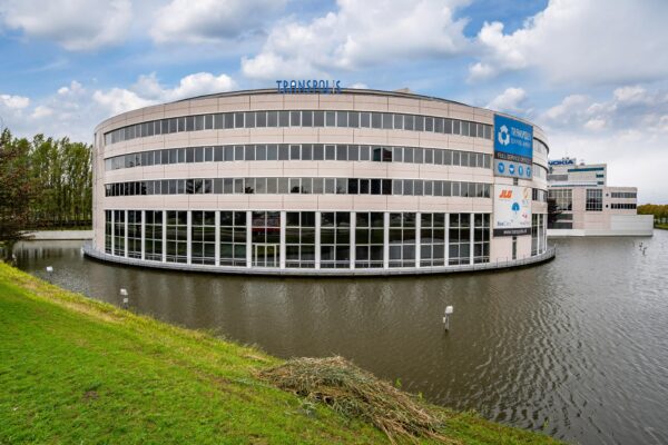 Een modern kantoorgebouw genaamd Transpolis, omgeven door water en een grasveld.